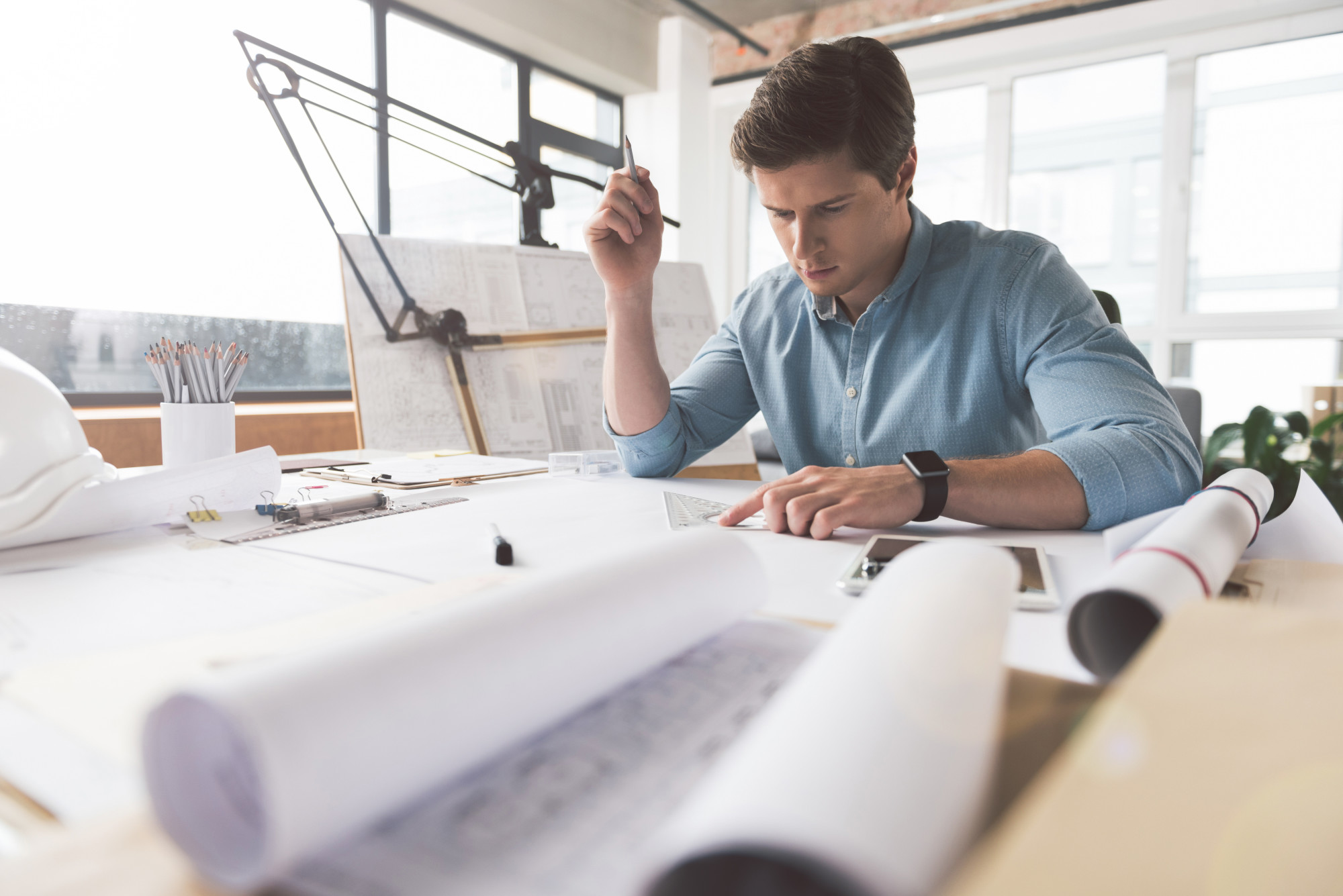 Involved in project. Serious qualified young engineer is drawing blueprint. He is sitting at desk and holding pencil while looking at paper thoughtfully. Copy space in the left side